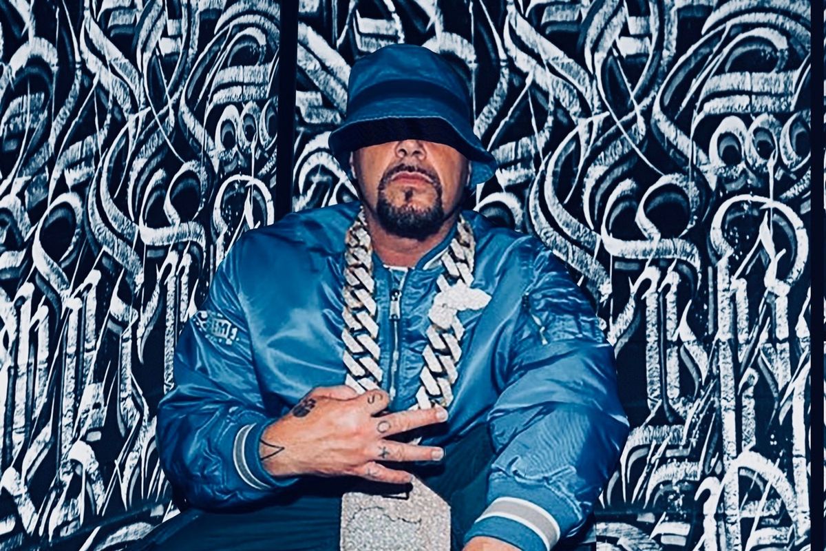 DJ Muggs poses for a photo in a blue jacket and blue hat, with a large chain. 