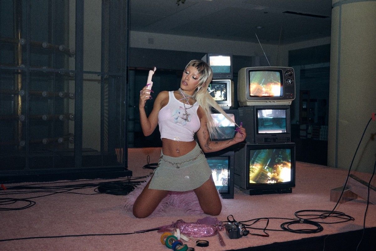 Doja Cat with blonde wig in front of TVs
