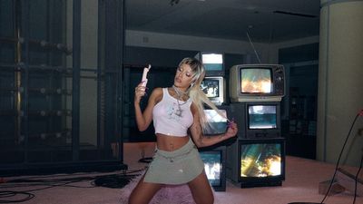 Doja Cat with blonde wig in front of TVs