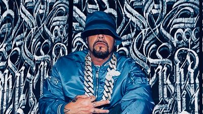 DJ Muggs poses for a photo in a blue jacket and blue hat, with a large chain. 