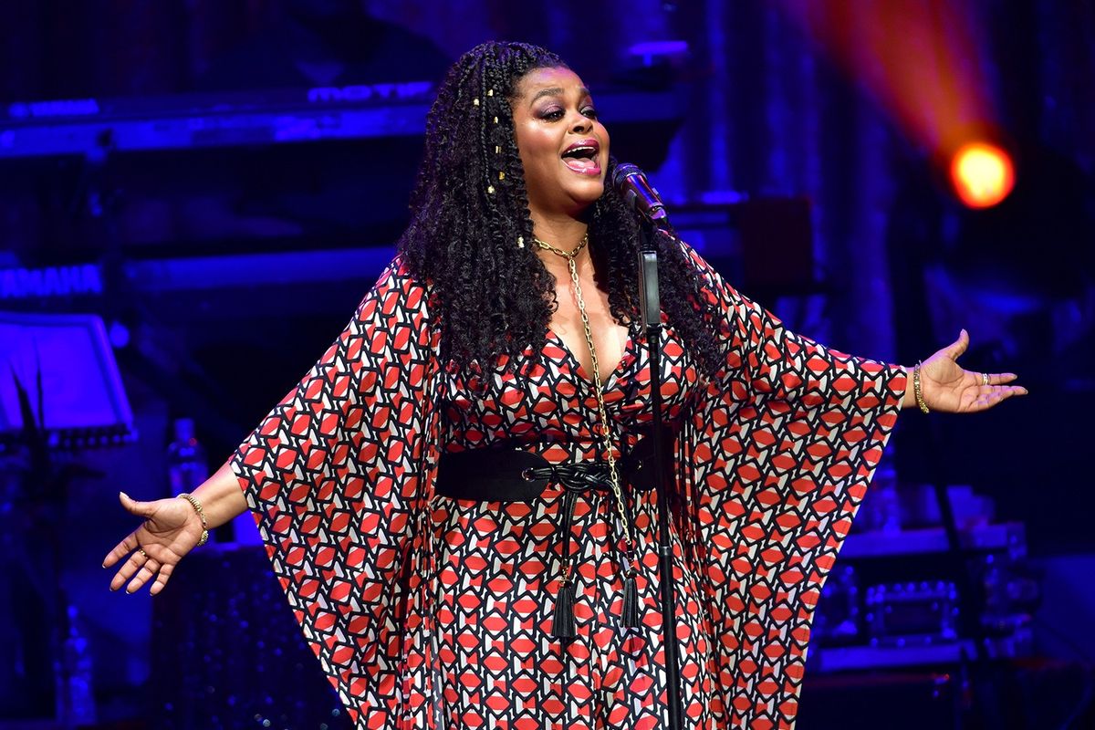 Jill Scott stands on stage singing into the mic with her arms wide