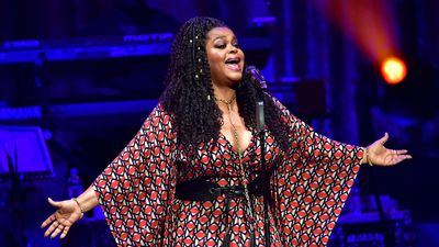 Jill Scott stands on stage singing into the mic with her arms wide