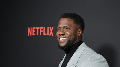 Kevin Hart in grey suit with black turtleneck smiling in front of Netflix movie premiere background
