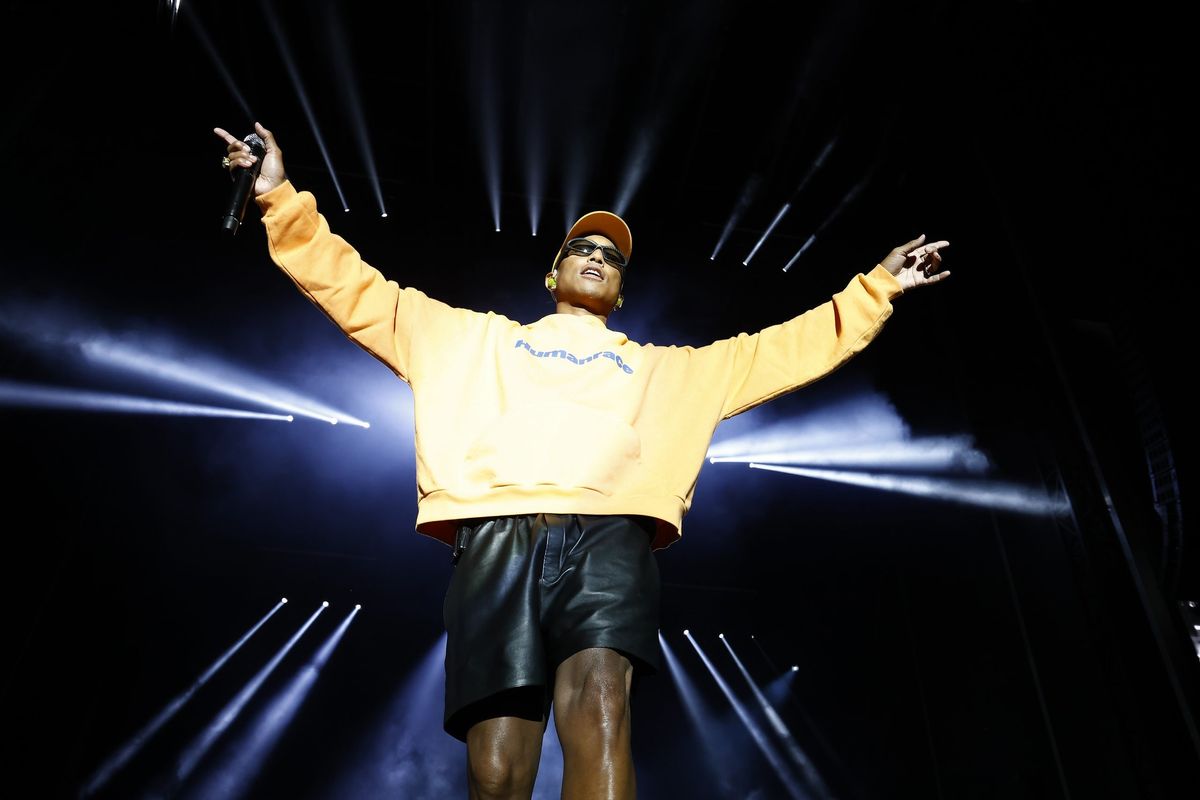 Pharrell on stage wearing a yellow crew neck and Black leather shorts performing with his arms out