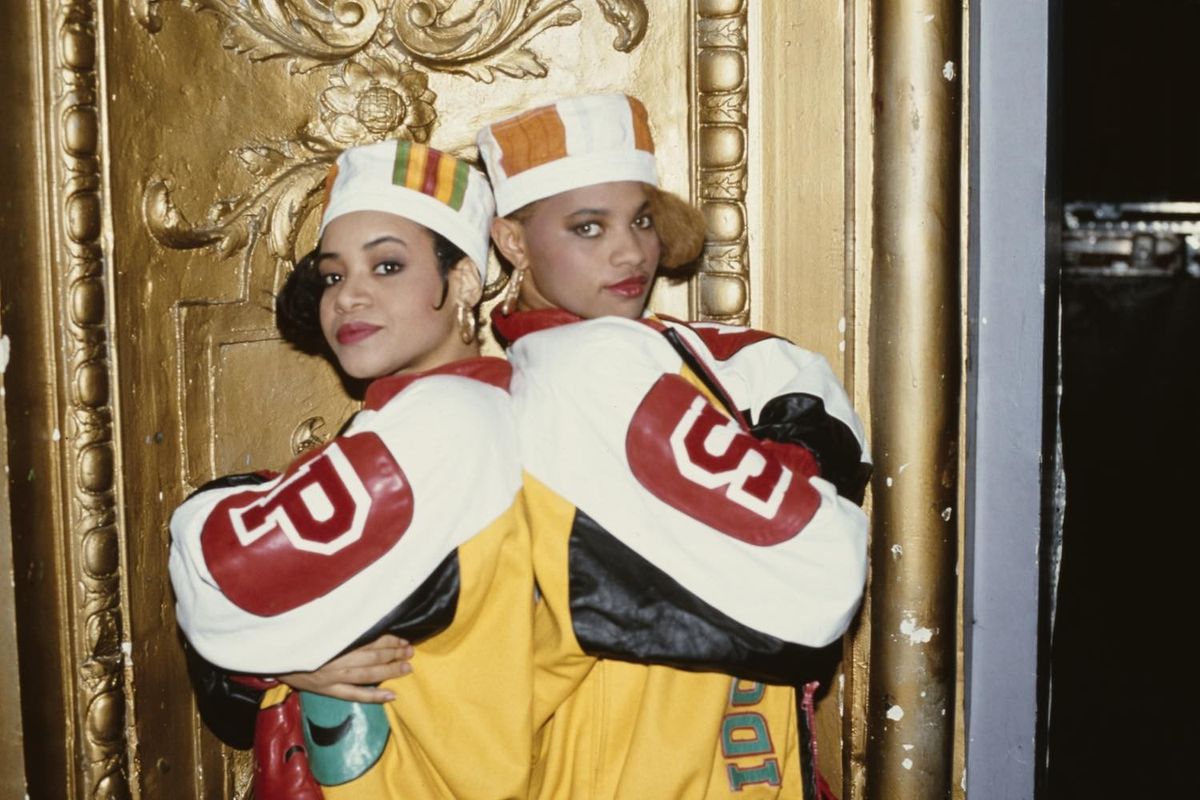 Salt-n-Pepa both wearing kente kufi hats and leather baseball jackets, posing back-to-back, both with their arms folded, United States, 1988. 