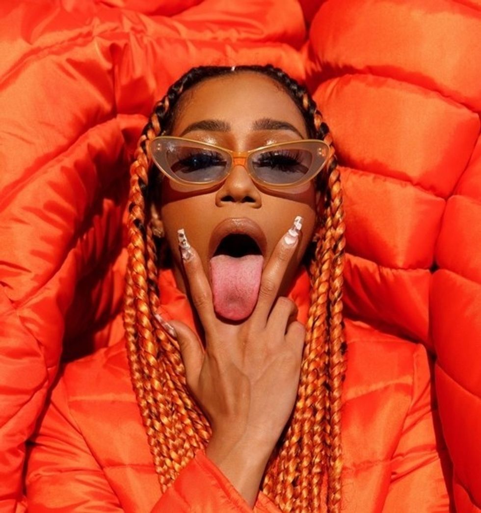 Boston rapper, BIA, in orange braids and puffy jacket making the v sign with tongue out