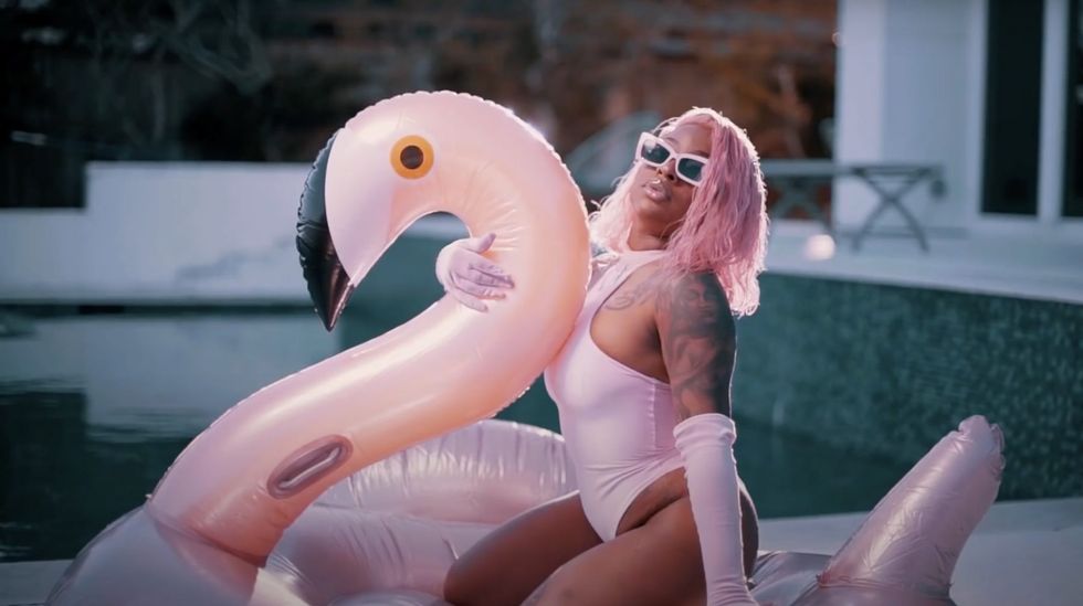 Female rapper with pink hair and pink one-piece swimsuit in the pool with flamingo float.