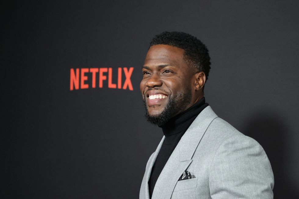 Kevin Hart in grey suit with black turtleneck smiling in front of Netflix movie premiere background