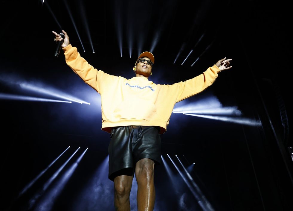 Pharrell on stage wearing a yellow crew neck and Black leather shorts performing with his arms out